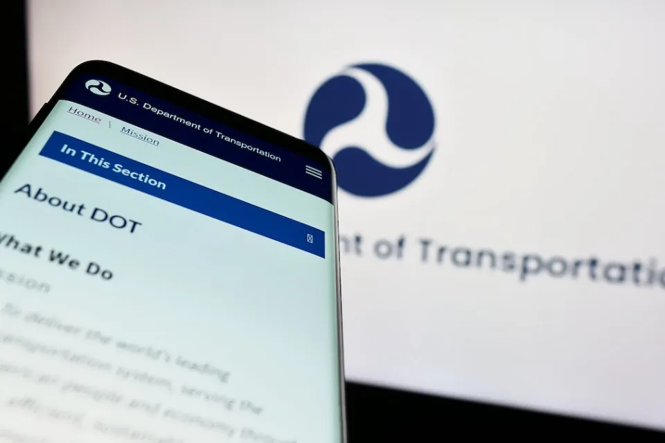 Close-up of smartphone with USDOT website pulled up and screen with USDOT logo in background.